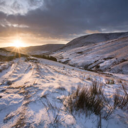 A winter dawn seen from the Pennine Way footpath, paused on the way up to Kinder Scout.  The sun rises over Rushup Edge, flooding the snow covered landscape with gentle light.   The footpath, opened in 1965, was the first long-distance footpath in the country.